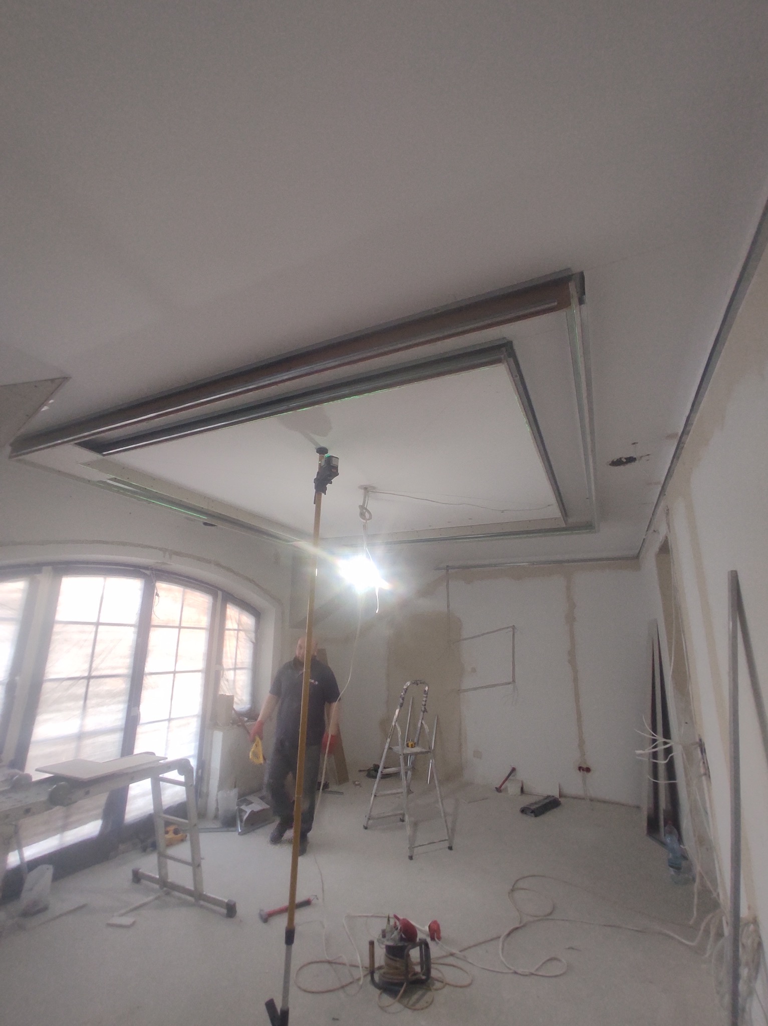 Wnętrze w trakcie prac wykończeniowych: widoczny sufit z metalową konstrukcją podwieszaną, ściany przygotowane do malowania, drabina, narzędzia i pracownik w tle.