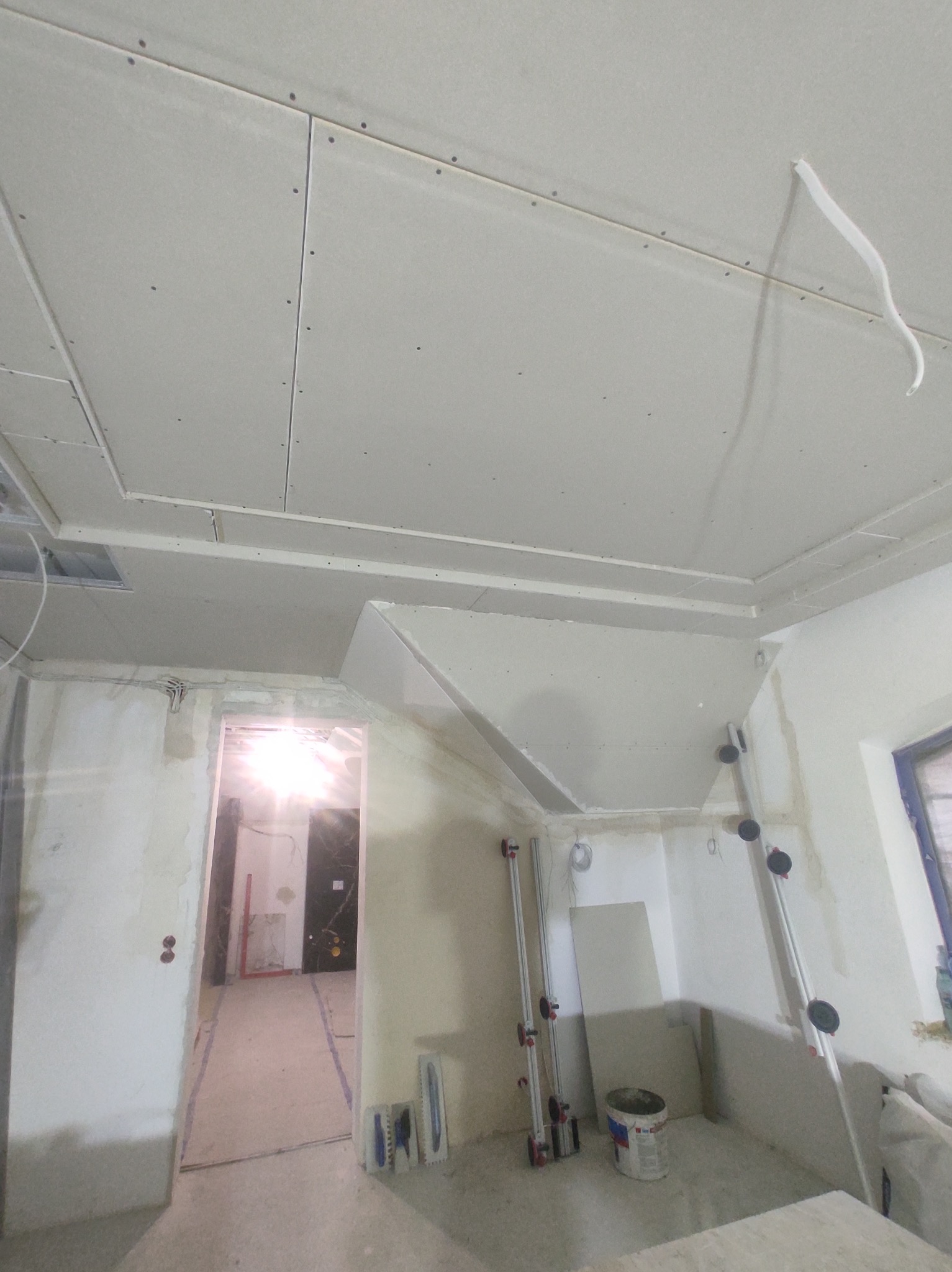 Sufit z płyt gipsowo-kartonowych z widocznymi łączeniami i wkrętami, fragment instalacji elektrycznej, drabiny z przyssawkami, wiadro farby i narzędzia do szpachlowania w pomieszczeniu w trakcie...