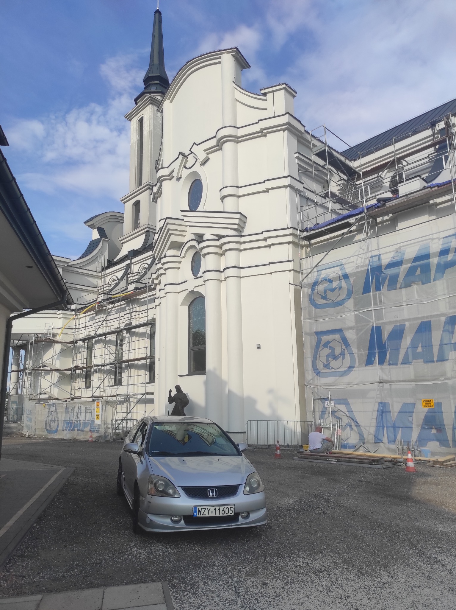 Biały kościół w trakcie remontu, widoczne rusztowania i siatka ochronna z niebieskim logo, na pierwszym planie srebrny samochód osobowy.
