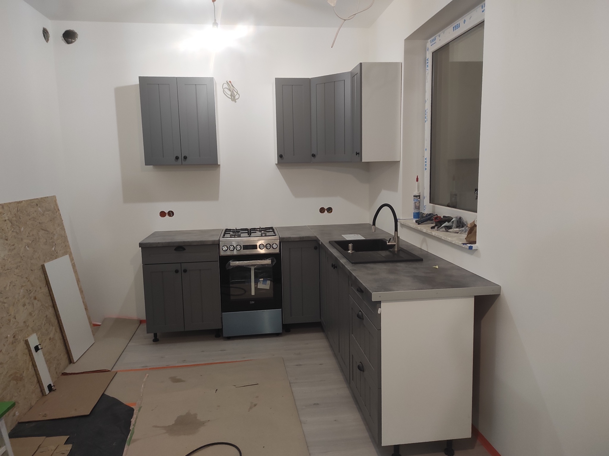 Szare szafki kuchenne z czarnymi uchwytami, zamontowane w surowym stanie pomieszczenia, widoczne ślady prac wykończeniowych na podłodze i niezabezpieczone kable.