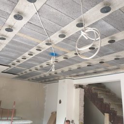 Proprofesional - Montaż izolacji akustycznej na suficie z widocznymi elementami mocującymi, instalacją elektryczną i fragmentem klatki schodowej w surowym stanie.