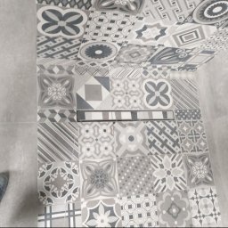 Proprofesional - Wykończona łazienka z patchworkową mozaiką szaro-białych płytek na podłodze i ścianie, z widocznym odpływem liniowym i fragmentem niebieskiego buta.