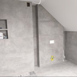 Proprofesional - Wykończona łazienka na poddaszu z szarymi, imitującymi beton płytkami ściennymi i podłogowymi; widoczna wnęka z akcesoriami i preparatem Sopro, instalacja podtynkowa WC oraz fragment dekoracyjnej...