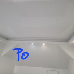 Proprofesional - Biały, błyszczący sufit napinany z oświetleniem punktowym, widok z dołu na skosach poddasza, z dopisanym niebieskim napisem 'Po' oraz fragmentem sylwetki osoby z boku.
