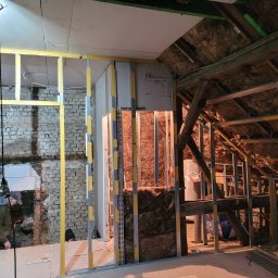 Proprofesional - Adaptacja poddasza: metalowa konstrukcja ścian działowych z izolacją z wełny mineralnej, fragment odsłoniętej ceglanej ściany, widoczne elementy instalacji elektrycznej i schemat na płycie gipsowej.