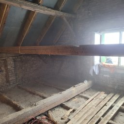 Proprofesional - Poddasze w trakcie remontu, widoczne ceglane ściany, drewniane belki stropowe i resztki podłogi, okno z widoczną osobą w tle.