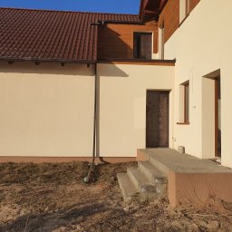 Proprofesional - Jasna elewacja domu jednorodzinnego z brązowym dachem, widoczne betonowe schody wejściowe i fragment ogrodu przed budynkiem.