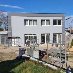 Proprofesional - Dwukondygnacyjny dom jednorodzinny w trakcie prac wykończeniowych elewacji, widoczni pracownicy, rusztowania oraz materiały budowlane na zewnątrz budynku.