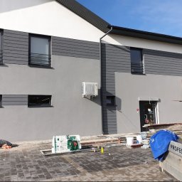 Proprofesional - Dwukondygnacyjny budynek po remoncie elewacji z szarym tynkiem i panelami elewacyjnymi, widoczny klimatyzator na ścianie oraz rusztowanie i materiały budowlane na pierwszym planie.