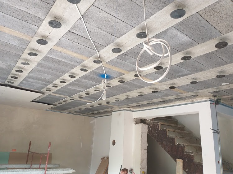 Montaż izolacji akustycznej na suficie z widocznymi elementami mocującymi, instalacją elektryczną i fragmentem klatki schodowej w surowym stanie.