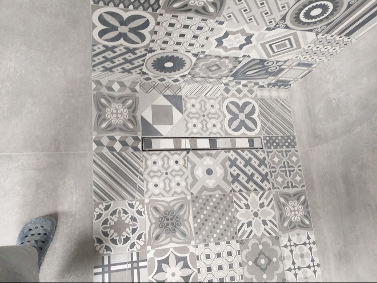 Wykończona łazienka z patchworkową mozaiką szaro-białych płytek na podłodze i ścianie, z widocznym odpływem liniowym i fragmentem niebieskiego buta.