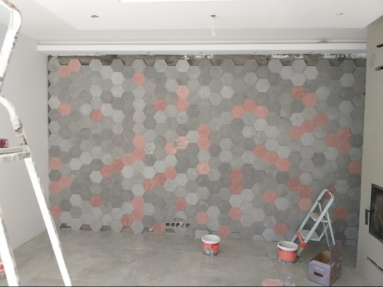Ściana w trakcie wykańczania z szarych i koralowych płytek heksagonalnych, widoczne puszki z farbą, drabina i niedokończone fugi