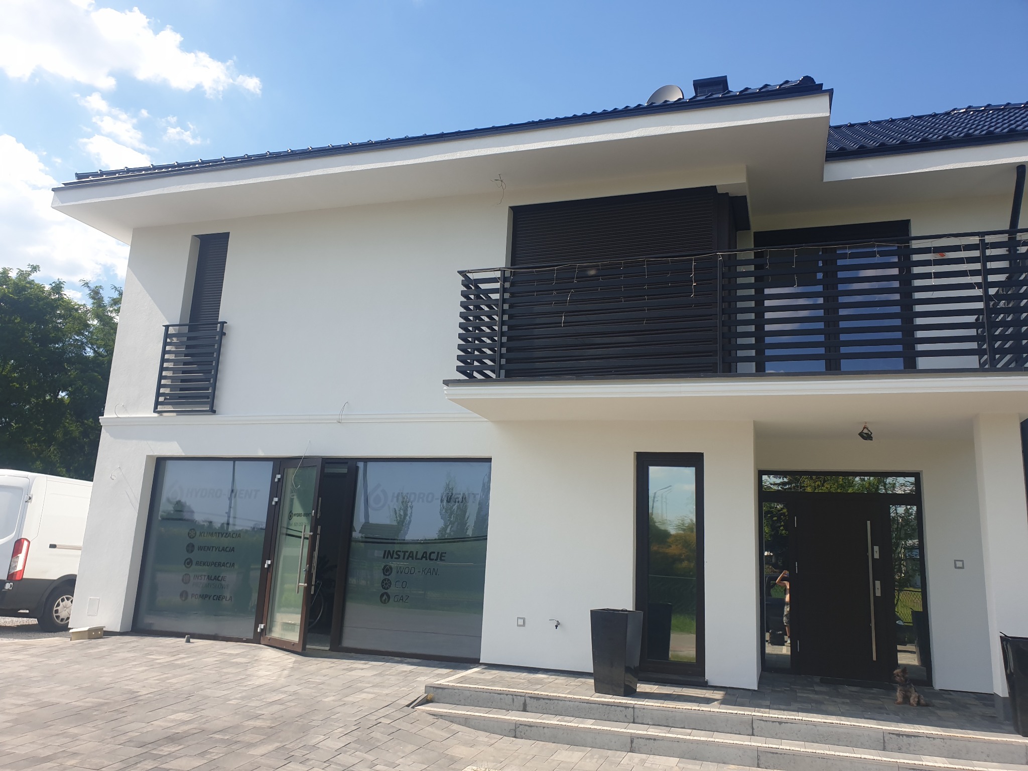 Nowoczesny, dwupiętrowy dom z białymi ścianami, czarnymi ramami okiennymi i balkonem z metalową balustradą, z widocznym logo firmy instalacyjnej na szklanych drzwiach wejściowych i zaparkowanym...