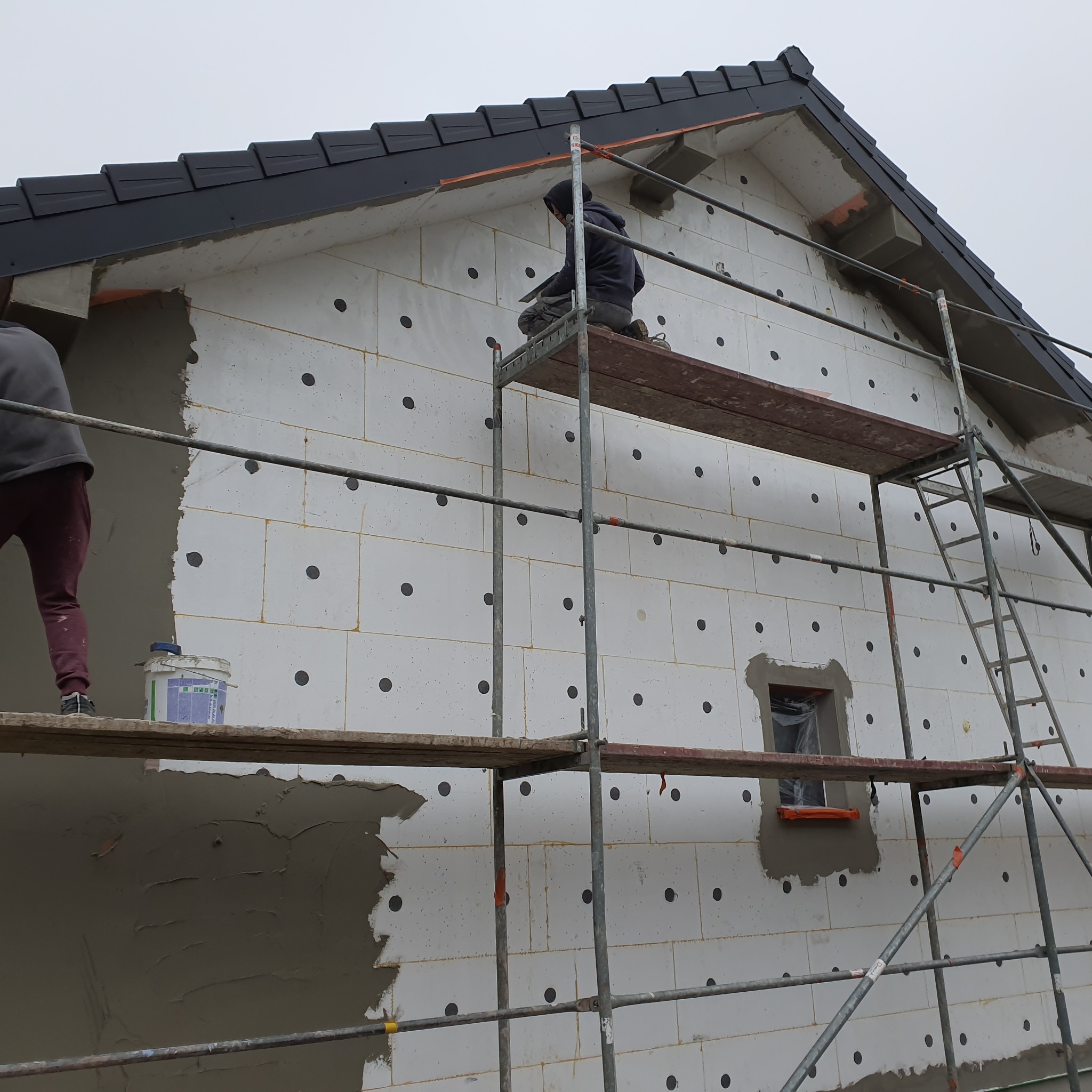 Elewacja domu w trakcie ocieplania styropianem z czarnymi kołkami, fragment ściany z nałożonym szarym tynkiem, pracownicy na rusztowaniu, pochmurne niebo.