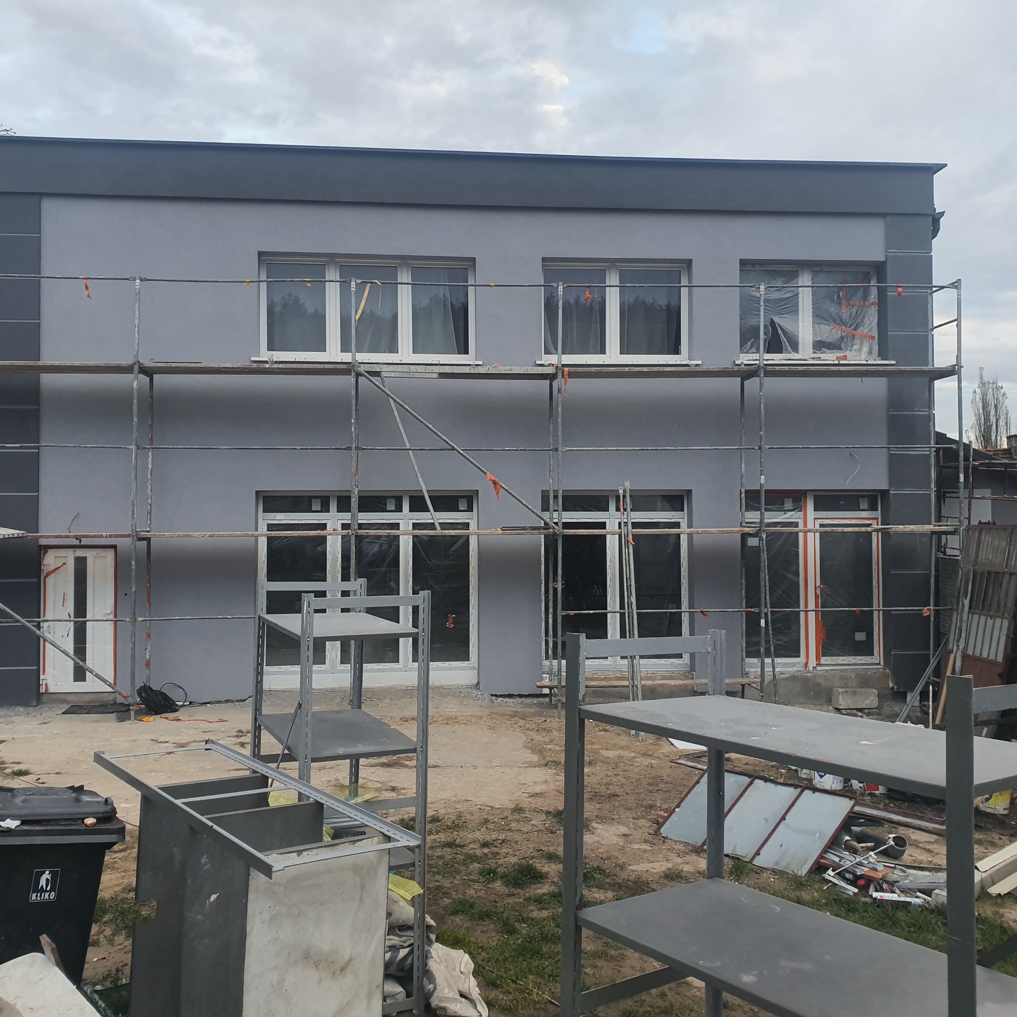 Szara elewacja dwukondygnacyjnego domu w trakcie malowania, widoczne rusztowania i zabezpieczone okna.