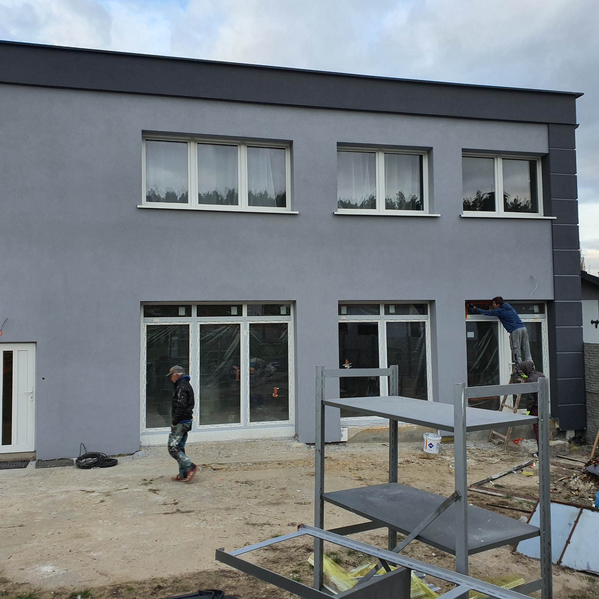 Dwupiętrowy budynek z szarą elewacją w trakcie malowania, pracownik na drabinie maluje ramę okienną, drugi przechodzi obok budynku, widoczne rusztowania i przygotowania do prac.