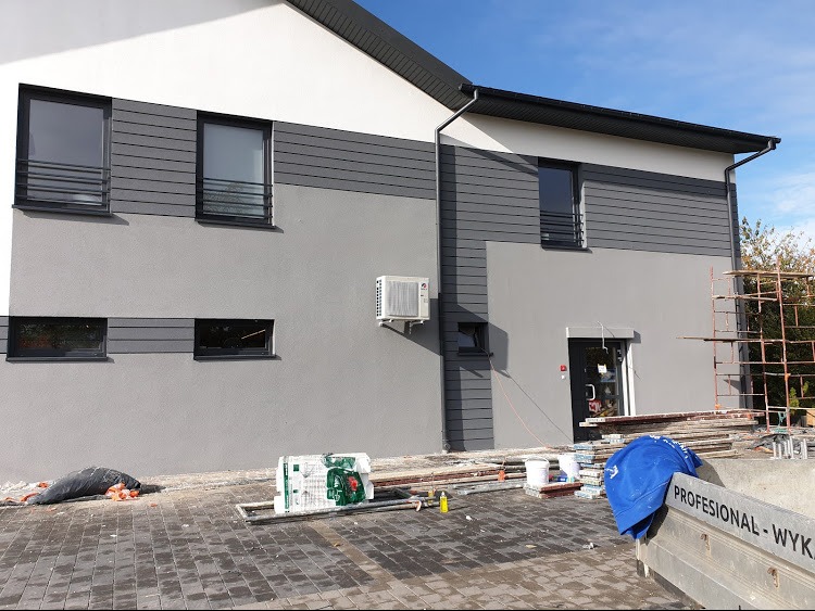 Dwukondygnacyjny budynek po remoncie elewacji z szarym tynkiem i panelami elewacyjnymi, widoczny klimatyzator na ścianie oraz rusztowanie i materiały budowlane na pierwszym planie.