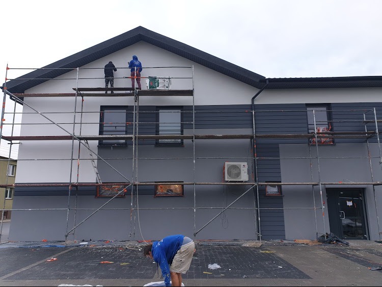 Dwóch pracowników na rusztowaniu podczas malowania elewacji budynku na szaro i biało, widoczny klimatyzator na ścianie.