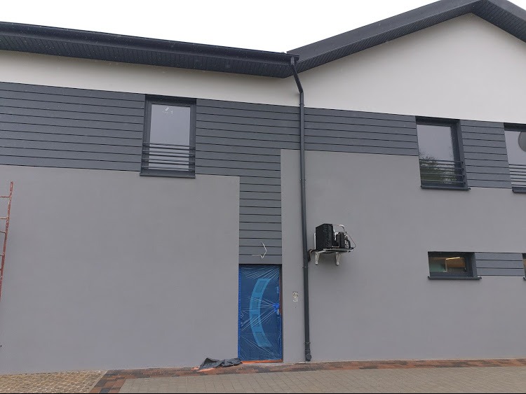 Dwukondygnacyjny budynek z szarą elewacją, częściowo wykończoną panelami imitującymi drewno, z klimatyzatorem zamontowanym na ścianie i drabiną opartą o ścianę po lewej stronie.