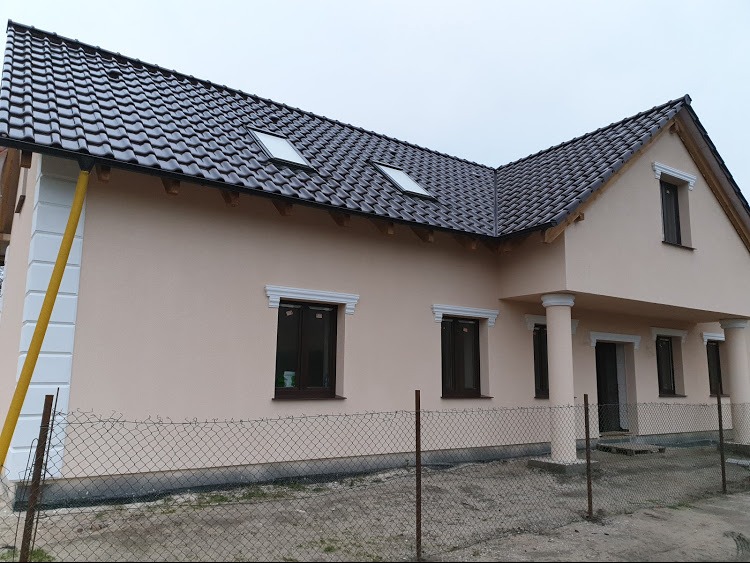 Jasnoróżowa elewacja domu jednorodzinnego z ciemnobrązowym dachem, ozdobnymi białymi elementami architektonicznymi wokół okien i na narożnikach, widok zza ogrodzenia z siatki.