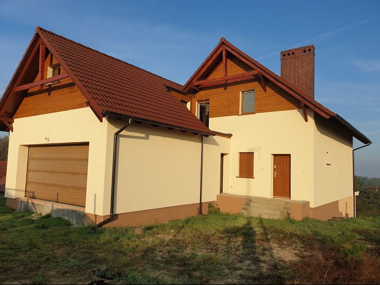 Dwukondygnacyjny dom z jasną elewacją i brązowym dachem, widoczny garaż z bramą rolowaną i komin z cegły klinkierowej, na nierównym terenie.