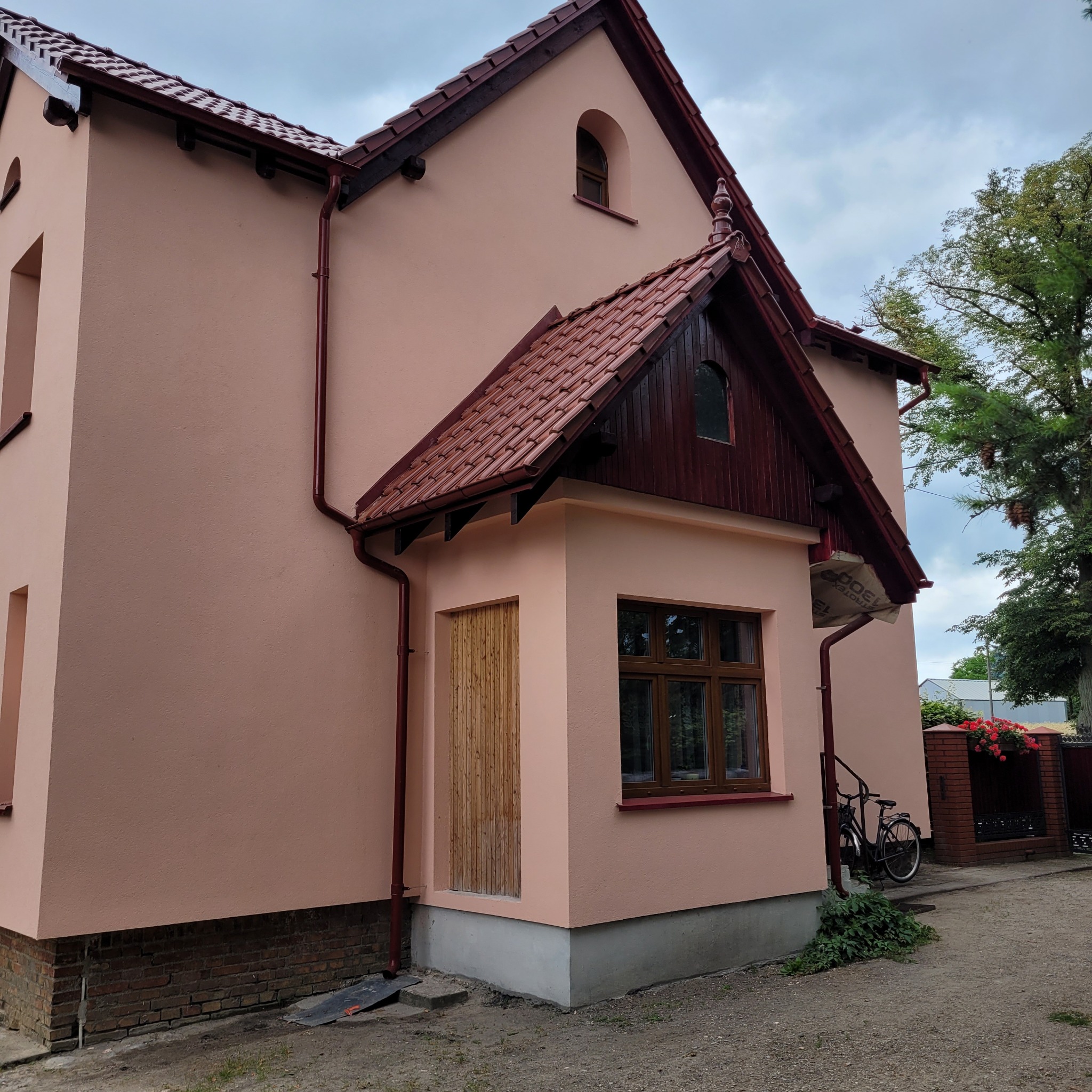Odnowiona elewacja domu z ciemnoczerwoną dachówką i rynnami, widoczna nowa, drewniana obudowa wejścia i okno, oraz rower oparty o ścianę.
