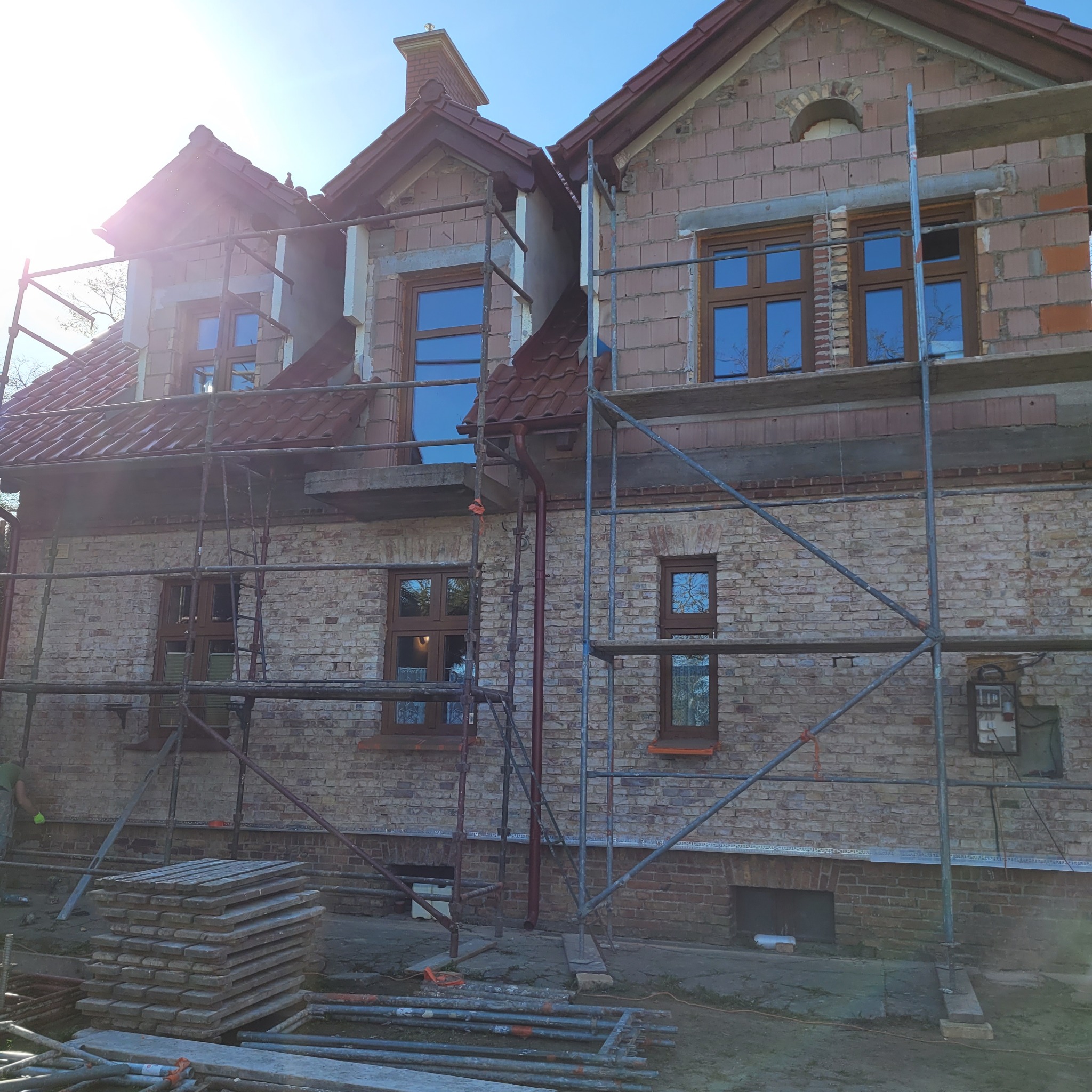 Dwupiętrowy dom z cegły w trakcie prac elewacyjnych, widoczne rusztowania, okna z brązowymi ramami i fragmenty ocieplenia.