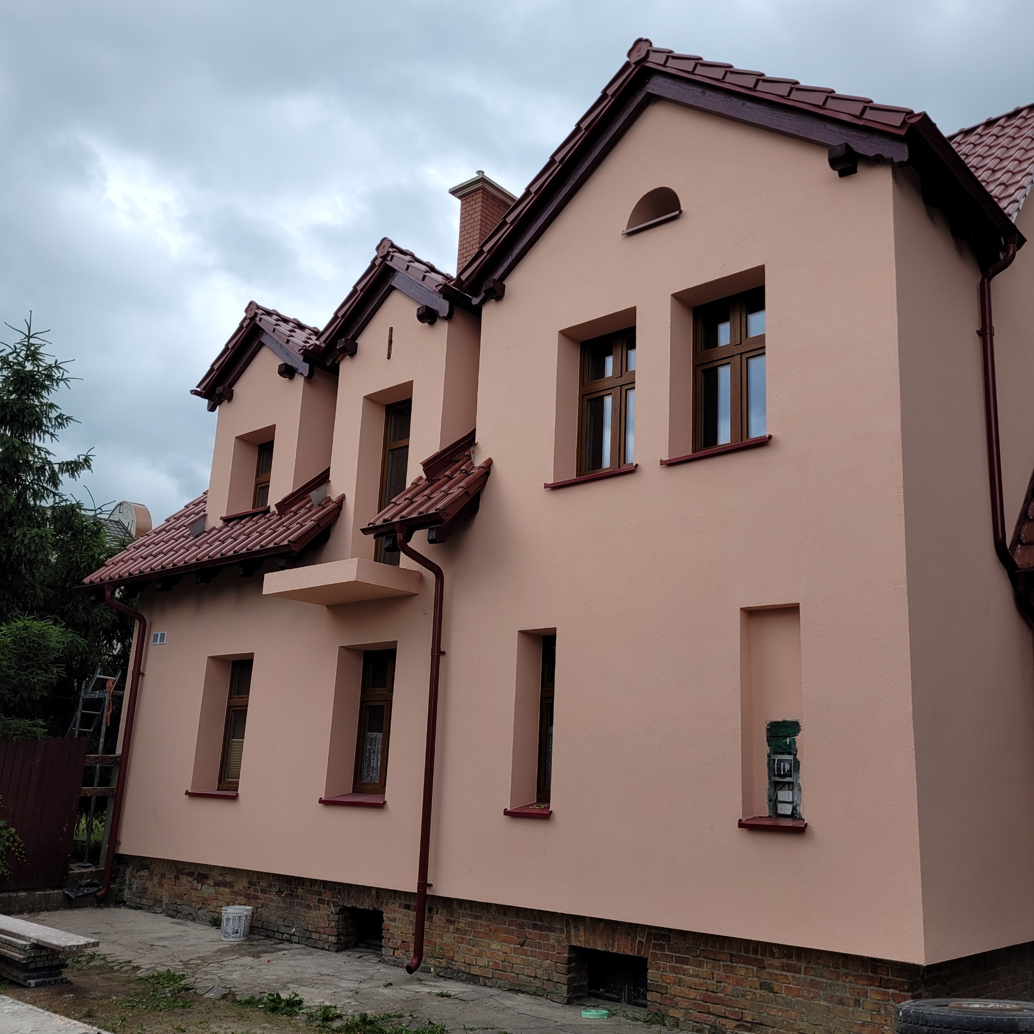 Dwupiętrowy dom z nową, łososiową elewacją, brązową stolarką okienną i dachem z czerwonej dachówki, widok z boku na tle pochmurnego nieba.