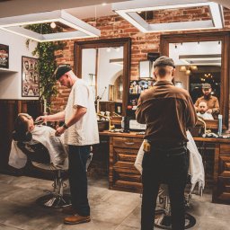 BarberHood Rzeszów - Wnętrze salonu: golibroda przy pracy, stylizacja brody brzytwą, drugi golibroda w tle, ceglana ściana, plakaty w ramach, oświetlenie LED.