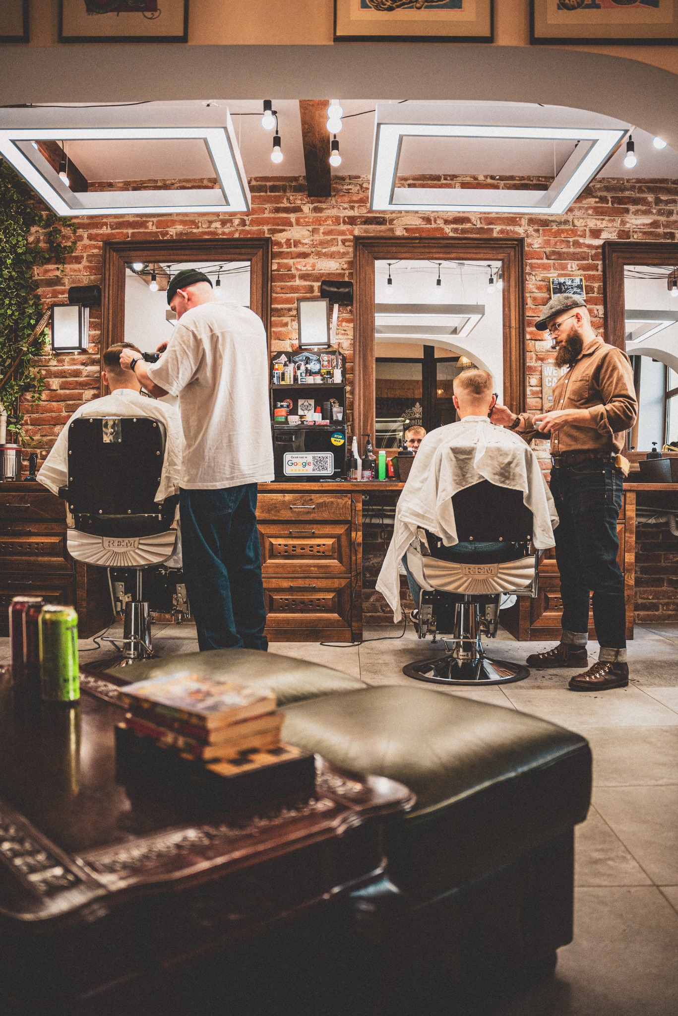 Fryzjerzy w trakcie pracy w salonie z ceglanymi ścianami, oświetleniem LED i klasycznymi fotelami fryzjerskimi.