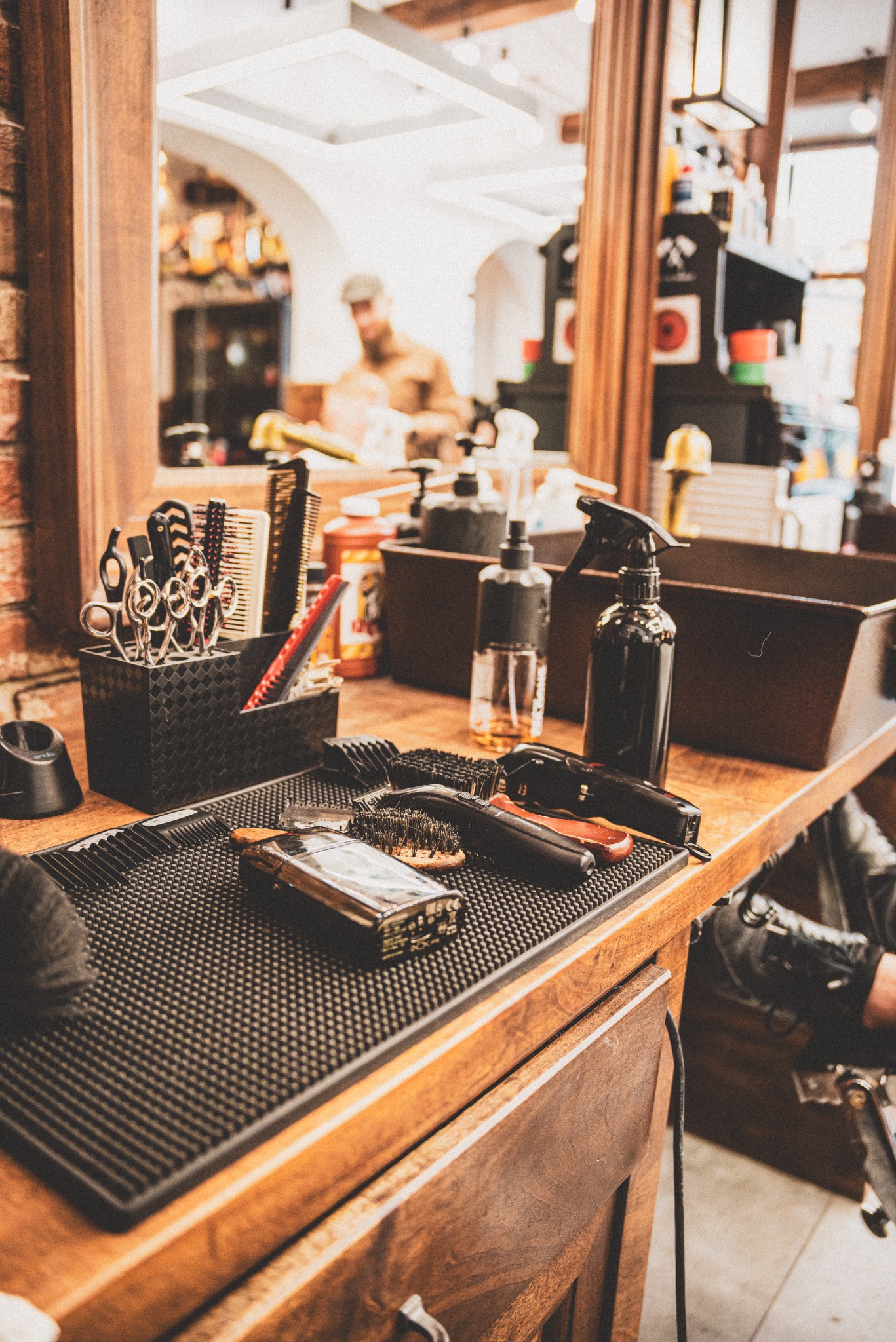 Przybornik z nożyczkami, grzebieniami, maszynkami i flakonami na drewnianym blacie w salonie, w tle widoczne lustro z odbiciem stylisty.