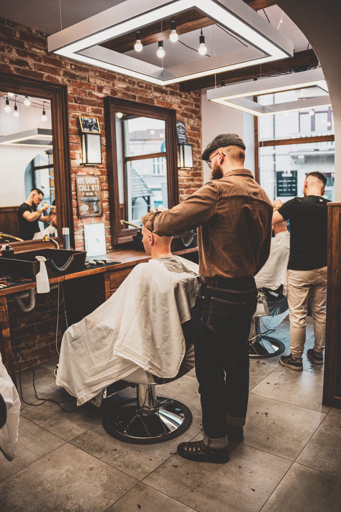 Mężczyzna w czapce strzyże klienta w salonie z ceglanymi ścianami i nowoczesnym oświetleniem, widoczne lustra i stanowiska pracy.