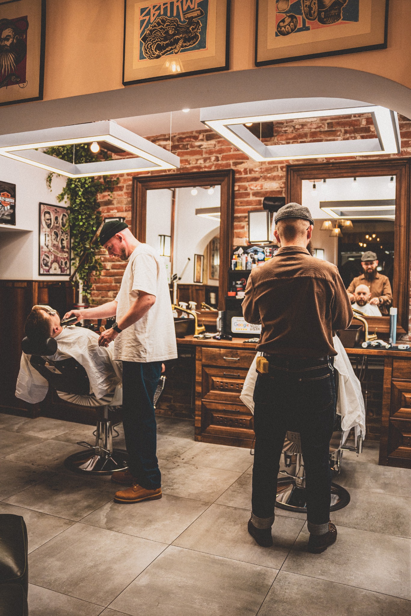 Wnętrze salonu: golibroda przy pracy, stylizacja brody brzytwą, drugi golibroda w tle, ceglana ściana, plakaty w ramach, oświetlenie LED.