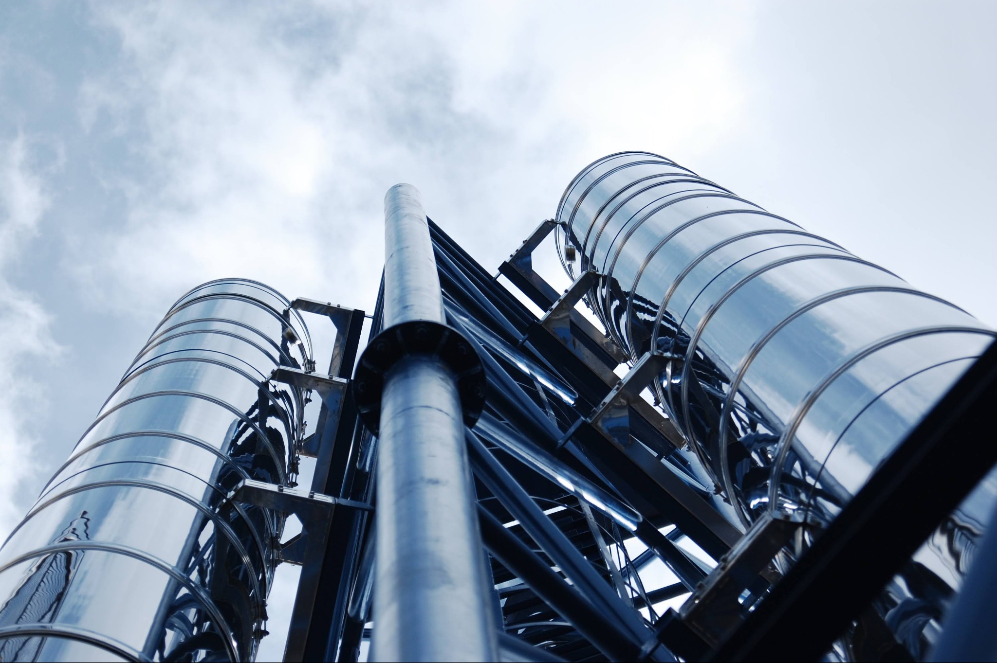 Zewnętrzna instalacja z metalowych rur, prawdopodobnie system wentylacyjny lub odprowadzania spalin, widziana z dołu na tle pochmurnego nieba.