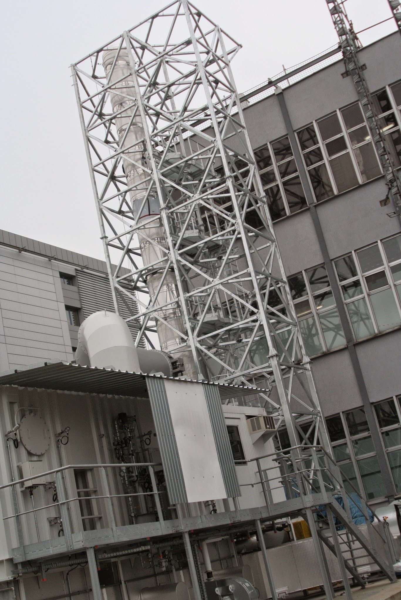 Zewnętrzna instalacja przemysłowa z metalową konstrukcją wsporczą, widoczne rury, zbiorniki i drabiny, na tle budynku z szarymi ścianami i oknami.