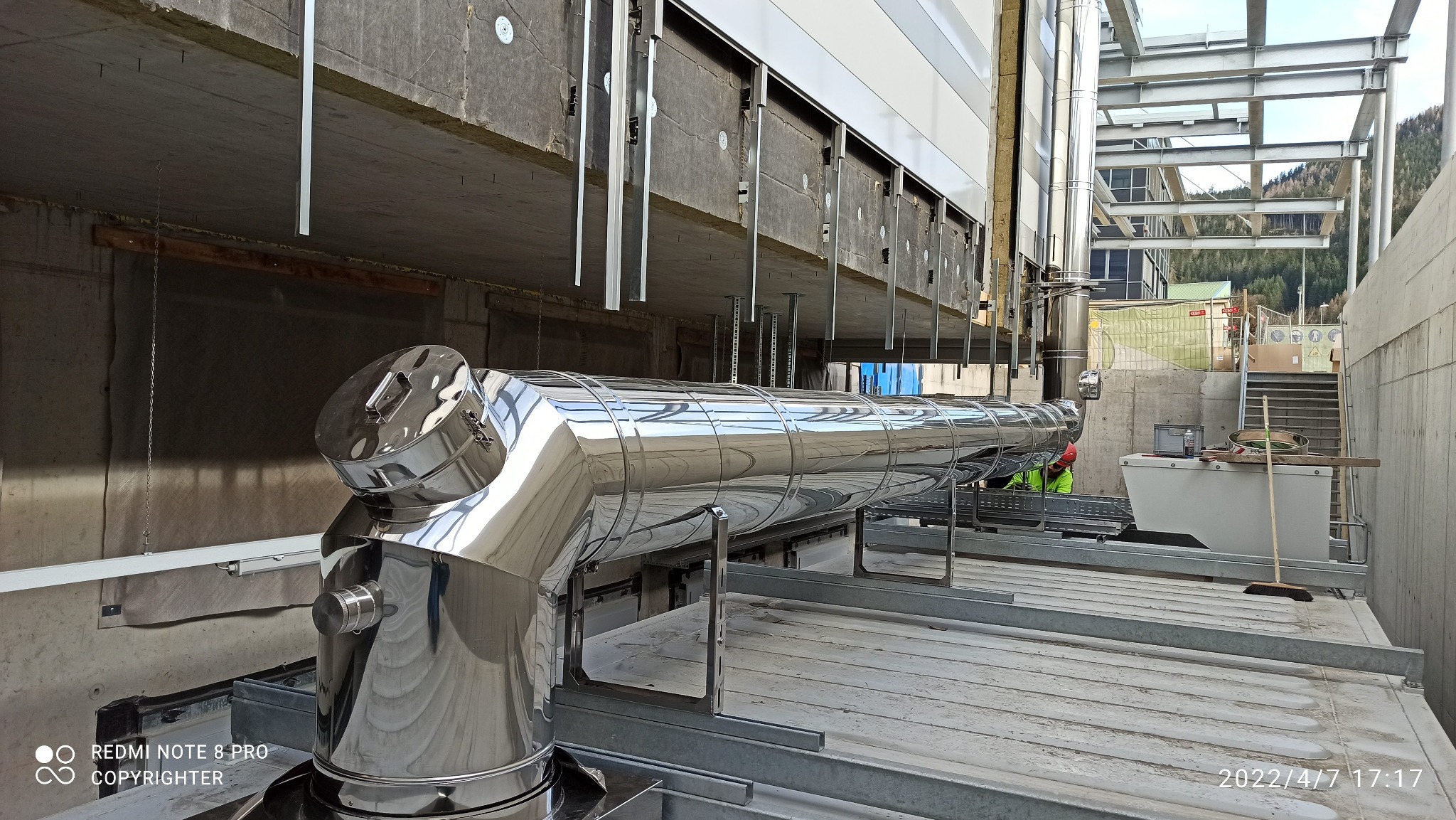 Błyszczący, metalowy przewód kominowy w trakcie montażu na dachu budynku, widoczny pracownik w odblaskowej kamizelce i czerwonym kasku.