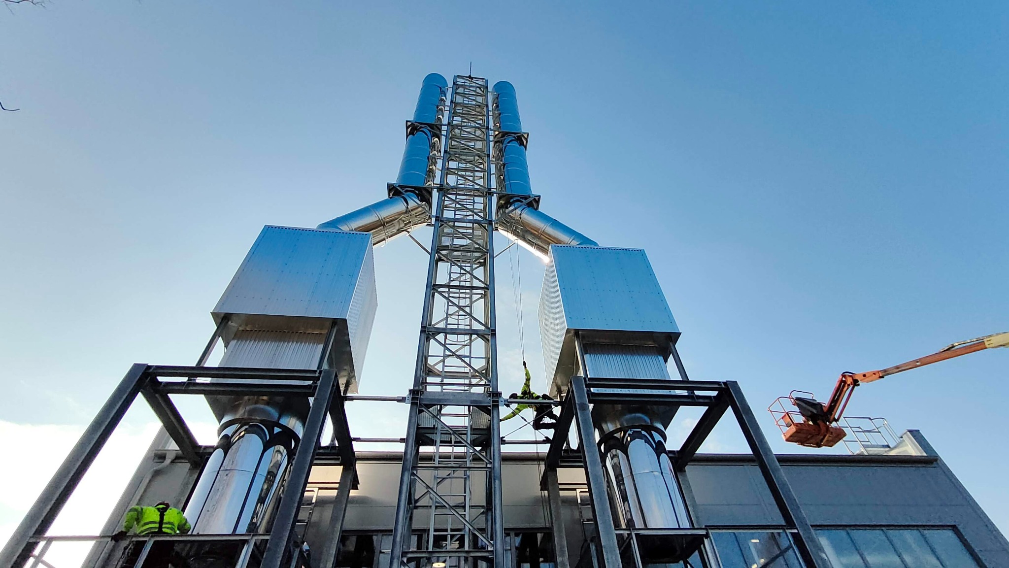 Montaż przemysłowego systemu odprowadzania spalin z dwoma pracownikami na rusztowaniu i podnośnikiem koszowym w słoneczny dzień.