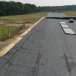 GJD SPÓŁKA Z OGRANICZONĄ ODPOWIEDZIALNOŚCIĄ - Widok z góry na czarny, płaski dach pokryty papą z widocznymi elementami wentylacyjnymi i obróbką blacharską na krawędzi, w tle drzewa i pole.