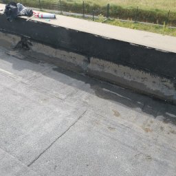 GJD SPÓŁKA Z OGRANICZONĄ ODPOWIEDZIALNOŚCIĄ - Widok z góry na fragment płaskiego dachu pokrytego papą, z widocznymi śladami napraw i narzędziami pracy (piana montażowa, szpachelka) leżącymi na krawędzi dachu, w tle pole i ogrodzenie.