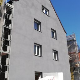 GJD SPÓŁKA Z OGRANICZONĄ ODPOWIEDZIALNOŚCIĄ - Budynek po remoncie elewacji, pomalowany na szaro, widoczne rusztowania i materiały izolacyjne, kobieta w okularach za ogrodzeniem.