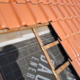 GJD SPÓŁKA Z OGRANICZONĄ ODPOWIEDZIALNOŚCIĄ - Fragment dachu z częściowo ułożoną czerwoną dachówką, widoczna membrana dachowa z nadrukiem. Elementy drewniane konstrukcji dachu. Ujęcie z góry, w słoneczny dzień.