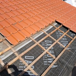 GJD SPÓŁKA Z OGRANICZONĄ ODPOWIEDZIALNOŚCIĄ - Częściowo ułożona, nowa, pomarańczowa dachówka ceramiczna na łatach i membranie dachowej. Widoczny fragment budynku w tle. Słoneczny dzień.