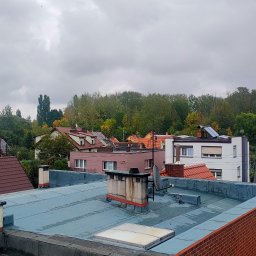 GJD SPÓŁKA Z OGRANICZONĄ ODPOWIEDZIALNOŚCIĄ - Widok na dachy budynków z płaskiego dachu pokrytego papą, kominy i anteny satelitarne na tle drzew i pochmurnego nieba. Widoczne panele słoneczne na jednym z budynków.