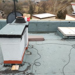 GJD SPÓŁKA Z OGRANICZONĄ ODPOWIEDZIALNOŚCIĄ - Płaski dach pokryty papą z kominem w trakcie renowacji. Widoczne narzędzia i materiały budowlane. W tle zabudowania i drzewa.