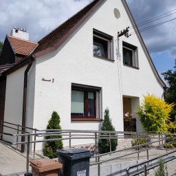 GJD SPÓŁKA Z OGRANICZONĄ ODPOWIEDZIALNOŚCIĄ - Biały dom z brązowym dachem i oknami, widoczny z zewnątrz, z rampą dla niepełnosprawnych i dwoma koszami na śmieci przed wejściem. Słoneczny dzień, błękitne niebo z chmurami.