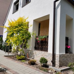 GJD SPÓŁKA Z OGRANICZONĄ ODPOWIEDZIALNOŚCIĄ - Biała elewacja domu z dekoracyjnym kamieniem, brązowymi rynnami i stolarką okienną. Żółte drzewko przy ścianie. Ujęcie z poziomu chodnika.