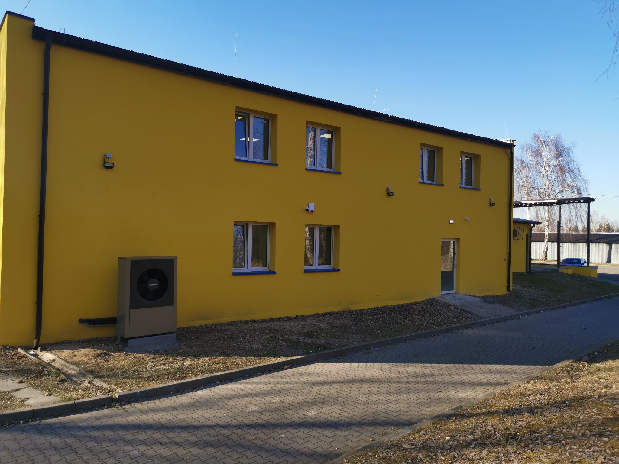 Żółty budynek z nową elewacją i pompą ciepła przy ścianie, widok z zewnątrz w słoneczny dzień.