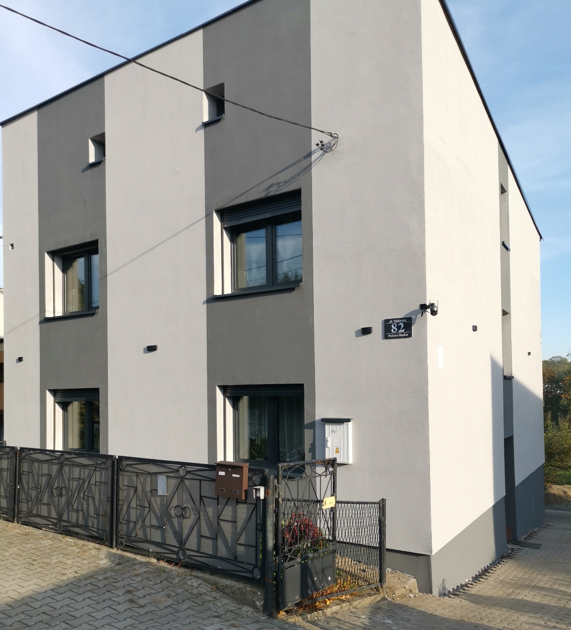 Dwukondygnacyjny dom z elewacją w odcieniach szarości, czarnymi ramami okiennymi i metalowym ogrodzeniem z dekoracyjnymi elementami.