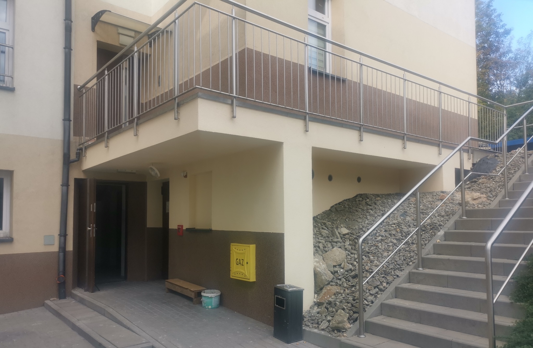 Nowoczesny budynek z balkonem z metalową balustradą i schodami zewnętrznymi z kamiennym nasypem. Widoczna skrzynka gazowa i wejście do budynku.