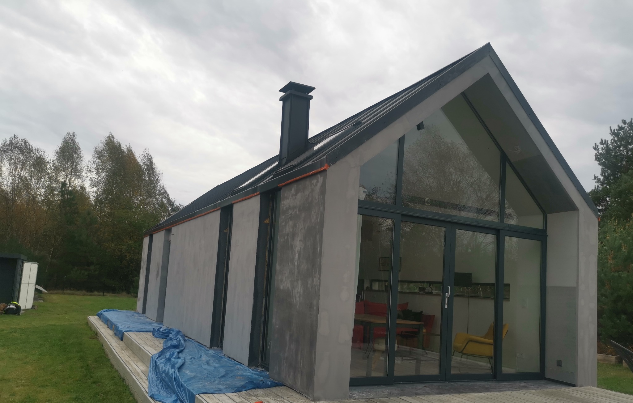 Nowoczesny dom jednorodzinny z szarą elewacją w trakcie prac wykończeniowych, widoczne zabezpieczające folie malarskie na tarasie, duże okna z ciemnymi ramami, czarny komin i dach.